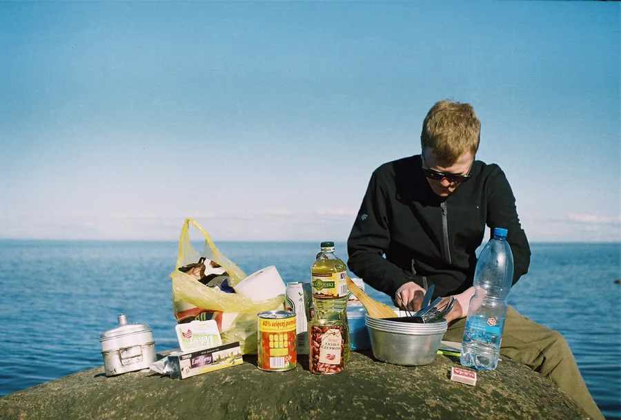 A man in sunglasses prepares food on a rock beside the sea.