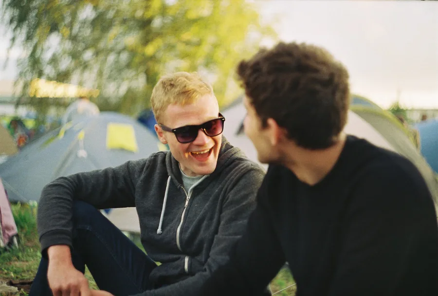 Two friends laugh together among tents at a campsite.