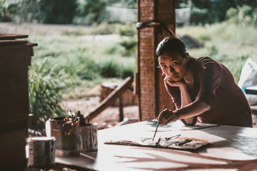 A woman pauses with a paintbrush at a sunlit work table.