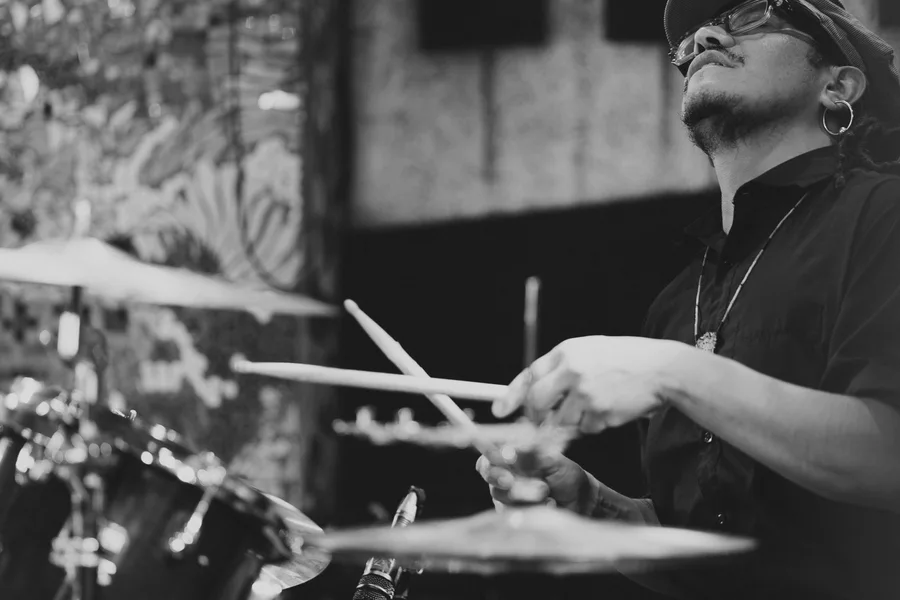 A drummer looks upward while playing, photographed in black and white.